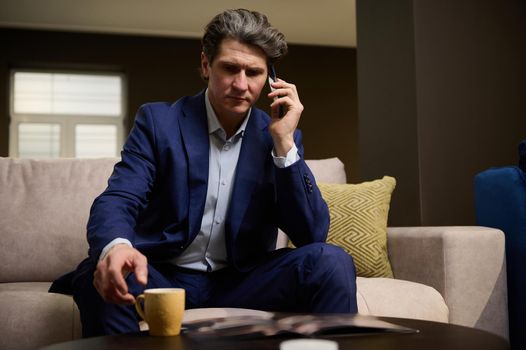 Portrait of a mature successful businessman talking on a mobile phone during a coffee break in his office. Handsome imposing entrepreneur, sales manager negotiating and advising clients on the phone