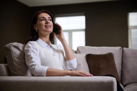 Attractive elegant successful European business woman, entrepreneur talking on mobile phone, sitting on a comfy velour sofa.