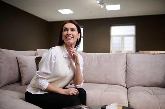 Charming brunette, elegant, middle-aged Caucasian woman smiles and looks away while sitting on a sofa in a furniture store. A cup of tea, a mobile phone and a catalog lie on a wooden coffee table