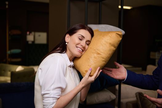 Beautiful dark-haired European woman buys pillows in a furniture store, puts her head on the cushion and smiles with pleasure. Concept of healthy sleep, interior design, home comfort and improvement