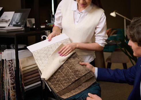 Close-up of a sales assistant showing fabric samples for sofa upholstery to customer in furniture store. Home decoration repair upholstery planning.