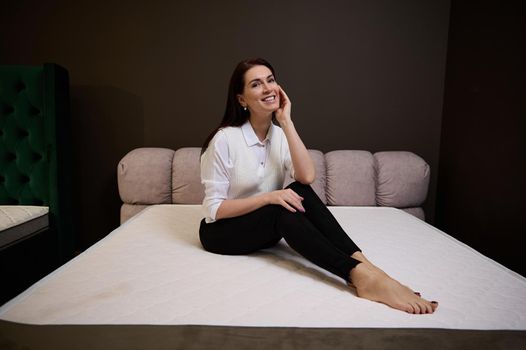 Attractive Caucasian pretty woman in casual attire sitting on an orthopedic mattress in the furniture store. The choice of mattresses in the exhibition center of a furniture store