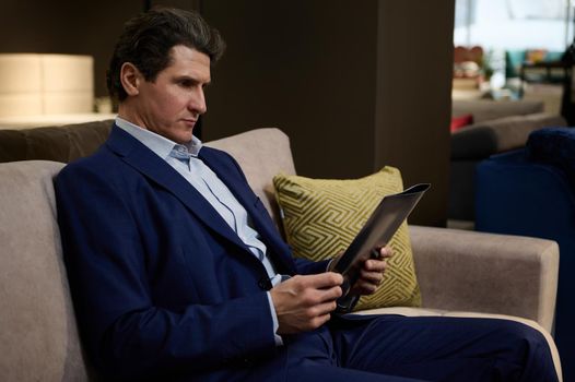 Confident mature Caucasian man sitting on a sofa in a furniture showroom and leafing a catalog with upholstered furniture