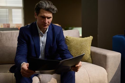 Handsome imposing mature Caucasian man in business suit looking at catalog with upholstered sofas and beds while visiting a furniture store showroom