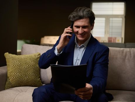 A charming successful businessman, a sales assistant in a furniture salon, holds a catalog in his hands and talks with a client on the phone, agreeing on the delivery time of the goods