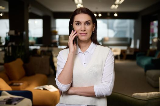 Beautiful confident female sales manager in furniture store talks on mobile phone looking at camera