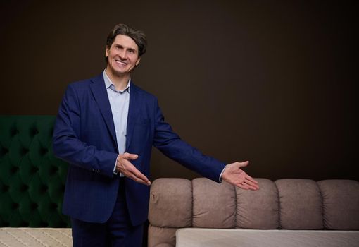 Handsome middle aged Caucasian man, sales manager, consultant, pointing at a stylish soft bed and orthopedic mattress in a furniture store showroom. Healthy sleep and good posture concept