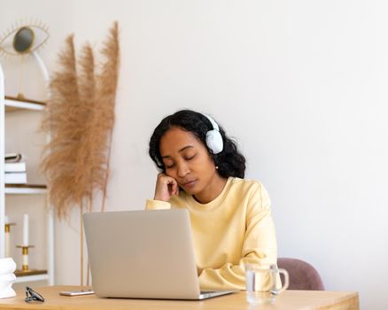 Bored and tired african-american woman student sleeping during online course on laptop at home. Exhausted millennial on quarantine. Workspace interior. Feeling lazy at lecture. Listening in headphones