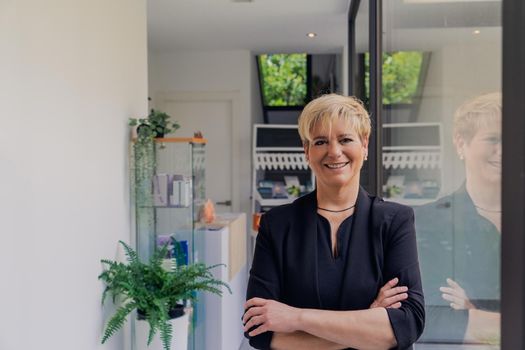 Corporate portrait smiling mature professional beautician with blonde hair, dressed in black work uniform, checking email on her tablet. owner in her small beauty salon looking at laptop. Work from home, enterprising woman, active small business owner. Relaxed atmosphere and soft lighting from window, natural light, tablet. horizontal.