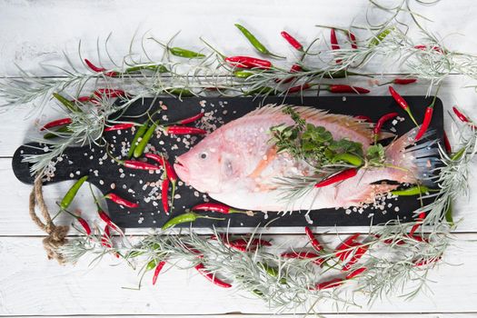 raw fresh pink tilapia fish lies on a board sprinkled with small red and green hot peppers, pink Himalayan salt and garnished with fresh curry sprigs, on white wooden planks. High quality photo