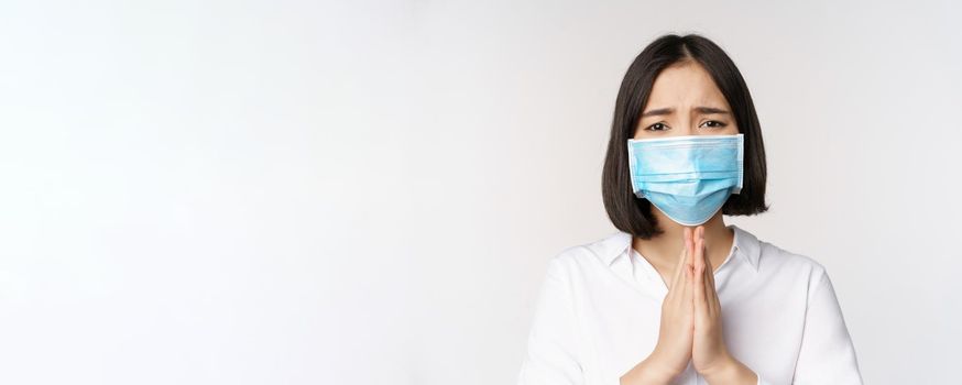 Portrait of asian woman in medical face mask from covid, begging, asking for help, say please, standing over white background.