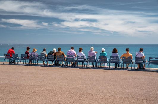 People sit on the famous blue chairs on embankment in Nice - France and watch at azure water of the Ligurian Sea, tranquillity, an old age, rest, age elderly people, the horizon, social. High quality photo