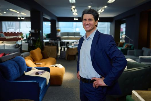 Confident business owner, entrepreneur, handsome imposing European mature man in the showroom of a furniture store with a wide range of sofas, couches and upholstered furniture