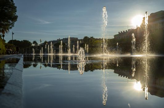 France, Nice, Famous place of Nice, square of fountain, mirror of water, playing kids, walking couples, a couple of old people are basking in the sunset, sun reflection, passing tram. High quality photo