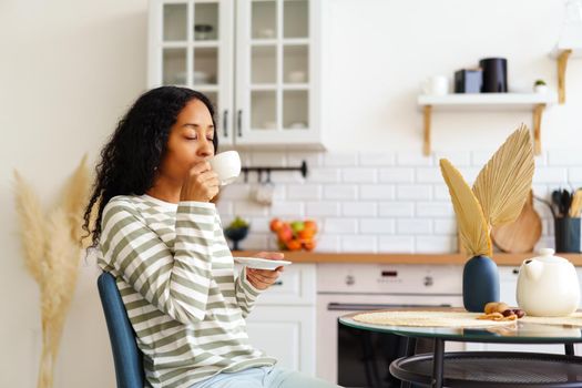 African-American woman drinking cup of coffee in morning for breakfast. Concept of taking time and slow-living niksen lifestyle. Modern white kitchen on background. Enjoying small break, micro-moment