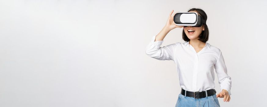 Excited young woman enter virtual reality in her glasses. Asian girl using vr headset, standing over white background.