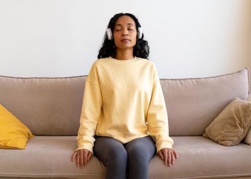 African-american woman having rest while sitting on sofa and listening to music in earphones. Concept of mindfulness and affirmative actions. Tranquil relax, meditation for personal fulfillment