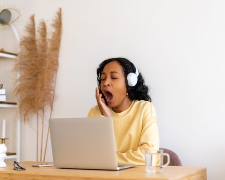 African-american lazy student yawning while listening to online course in earphones on laptop. Woman sitting at home and remote working. Millennial working extra hours and getting tired of workload