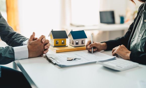 Real estate agent assisting client to sign contract at desk with house model.