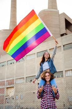 Young happy woman sitting on male friend shoulders waving rainbow flag in LGBT rights demonstration. Vertical image.