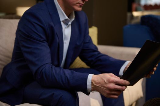 Hands of a mature man in business suit holding a catalog and sitting on a sofa in the showroom of an upholstered furniture store.