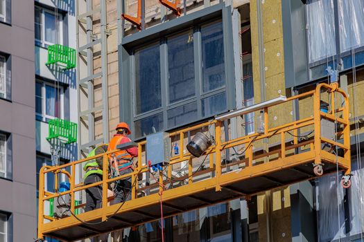 Male builders work on the facade of the building in a suspended cradle. Construction works.