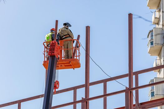 Male builders work at height in a lifting cradle, creating the iron frame of the building.
