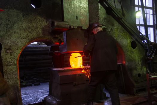 gray haired blacksmith worker forging large piece of hot steel in old factory with big steam hammer in Tula, Russia - February 1, 2020