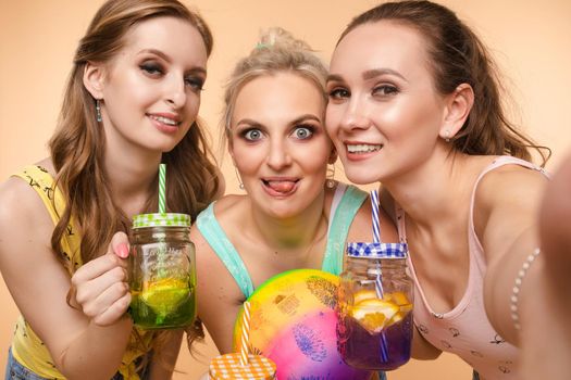 Three beautiful slender women in summer clothes with cocktails are laughing, having a good time together. Studio, isolated background. The concept of recreation