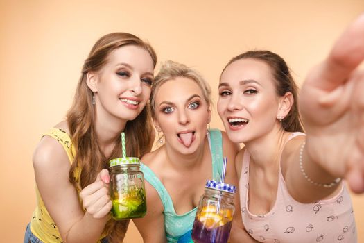 Three beautiful slender women in summer clothes with cocktails are laughing, having a good time together. Studio, isolated background. The concept of recreation