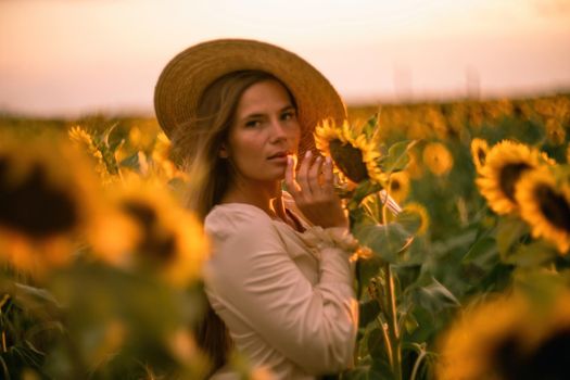 Beautiful middle aged woman looks good in a hat enjoying nature in a field of sunflowers at sunset. Summer. Attractive brunette with long healthy hair