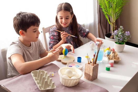Smiling little girl and boy are sitting at a white table, at home, by the window, painting eggs for the Easter holiday
