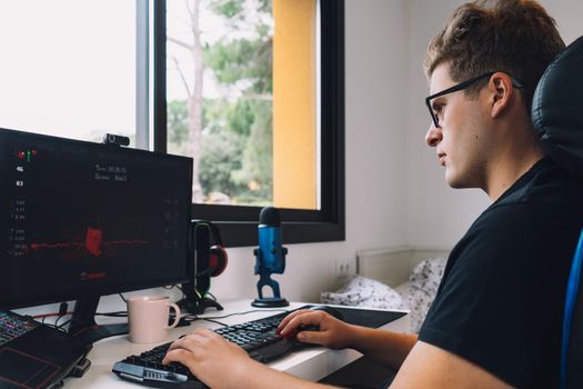 young man with short blond hair wearing glasses, concentrated and hard working, dressed in black shirt, sitting in a gamer chair, working as a graphic designer from home on his computer. natural light from window desk, with computer screen keyboard and coloured lights. Horizontal