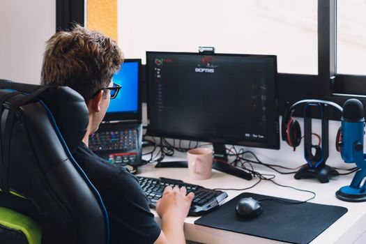 young man with short blond hair wearing glasses, concentrated and hard working, dressed in black shirt, young blond man working sitting in his desk chair at home at his computer. natural light from window desk, with computer screen keyboard and coloured lights. Horizontal