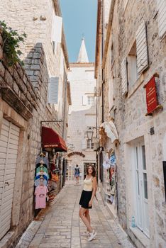 Girl tourist walking through ancient narrow street on a beautiful summer day in MEDITERRANEAN MEDIEVAL CITY , OLD TOWN bUDVA, MONTENEGRO. Young beautiful cheerful woman walking on old street at tropical town. Pretty girl looking at you and smiling