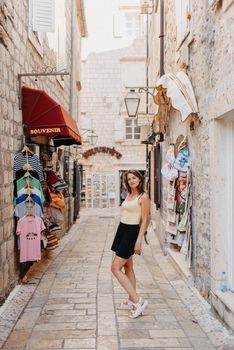 Girl tourist walking through ancient narrow street on a beautiful summer day in MEDITERRANEAN MEDIEVAL CITY , OLD TOWN bUDVA, MONTENEGRO. Young beautiful cheerful woman walking on old street at tropical town. Pretty girl looking at you and smiling