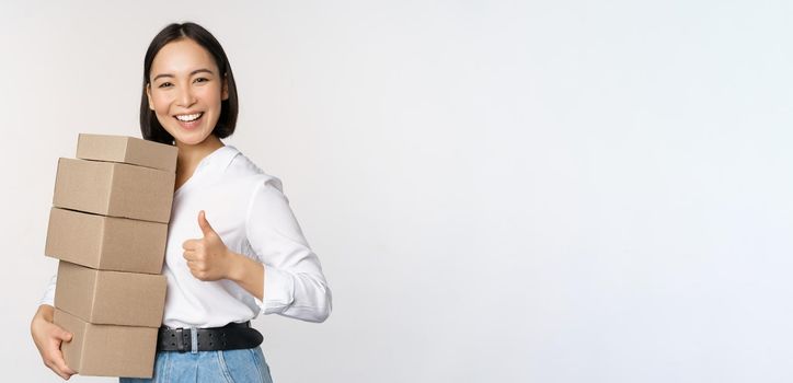 Image of happy modern asian woman showing thumbs up, holding boxes delivery goods, standing against white background.