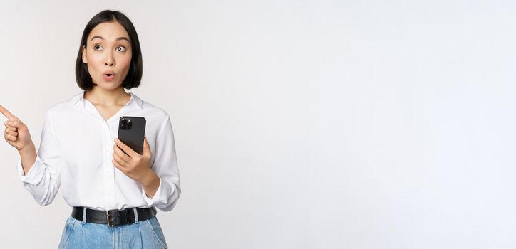 Excited asian girl shopping on mobile app, holding phone and pointing finger left at empty copy space, showing promo text, standing with smartphone against white background.