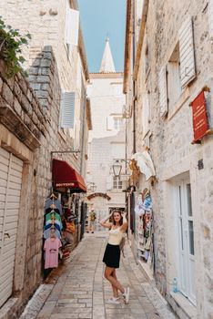 Girl tourist walking through ancient narrow street on a beautiful summer day in MEDITERRANEAN MEDIEVAL CITY , OLD TOWN bUDVA, MONTENEGRO. Young beautiful cheerful woman walking on old street at tropical town. Pretty girl looking at you and smiling