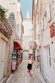 Girl tourist walking through ancient narrow street on a beautiful summer day in MEDITERRANEAN MEDIEVAL CITY , OLD TOWN bUDVA, MONTENEGRO. Young beautiful cheerful woman walking on old street at tropical town. Pretty girl looking at you and smiling