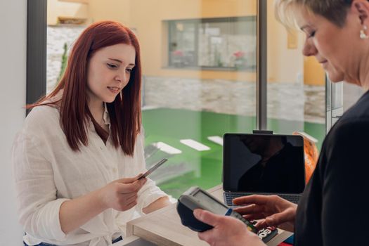 Mature beautician with smiling blonde hair, dressed in black work uniform, charging for a service at the counter of her shop. Young red-haired girl, dressed in white shirt and jeans, salon customer paying for a service with her credit card. Relaxed atmosphere and soft lighting from window, natural light, presentation brochures, horizontal.