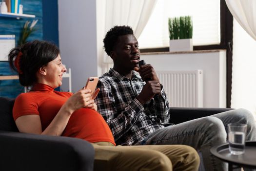 Pregnant caucasian woman laughing at african american man singing at remote control while sitting on sofa in living room. Joyful adult couple acting funny while expecting baby at home.