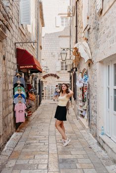 Girl tourist walking through ancient narrow street on a beautiful summer day in MEDITERRANEAN MEDIEVAL CITY , OLD TOWN bUDVA, MONTENEGRO. Young beautiful cheerful woman walking on old street at tropical town. Pretty girl looking at you and smiling