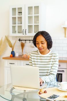 African-American woman working on laptop while sitting in dining room. Concentrated on remote work from home. Concept of telecommuting and teleworking in kitchen