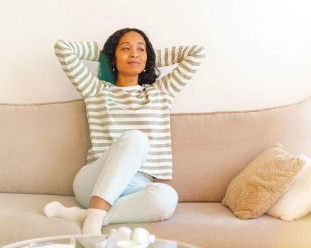 Happy African-American female throwing hands behind head while sitting on couch in living room. Concept of slow pace lifestyle. Showing positive emotion when having rest. Comfy clothes, home interior