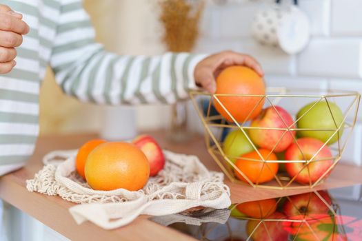 Unrecognizable person sorting out ripe apples and oranges in bowl after doing grocery with eco bag. Concept of healthy eating snack. Fresh and tasty food for vegetarians