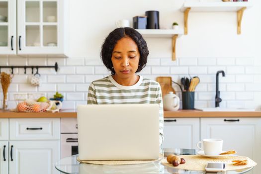 Concentrated ethnic woman using computer at table in kitchen. Busy female working from home. Remote workspace on quarantine. Concept of surfing the Net as leisure activity