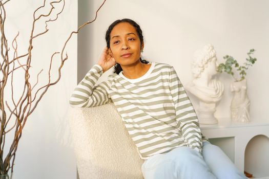 Calm and pensive African-American woman in casual clothing dreaming while looking at window in aesthetic lounge. Natural sunset light on background. Horizontal, copy space