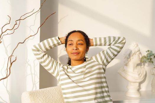 Calm and smiling African-American woman dreaming while sitting in lounge in front of window. Natural light from sunset. Concept of self-reflection and mindfulness. Morning time while enjoying life
