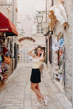 Girl tourist walking through ancient narrow street on a beautiful summer day in MEDITERRANEAN MEDIEVAL CITY , OLD TOWN bUDVA, MONTENEGRO. Young beautiful cheerful woman walking on old street at tropical town. Pretty girl looking at you and smiling